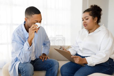 Photo for Mental health concept. Distressed upset young black man patient sitting on couch, holding napkin, crying during session with psychotherapist hispanic lady plus size, clinic interior, copy space - Royalty Free Image
