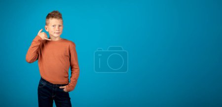 Photo for Call Me. Preteen Caucasian Boy With Hand Near Ear Imitating Phone And Looking At Camera, Tween Male Child Inviting For Communication While Standing On Blue Studio Background, Panorama - Royalty Free Image