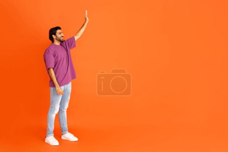 Foto de Alegre feliz guapo elegante millennial oriental chico en traje casual hipster ondeando en el espacio de copia y sonriendo, saludando a alguien, fondo de estudio naranja, longitud completa - Imagen libre de derechos