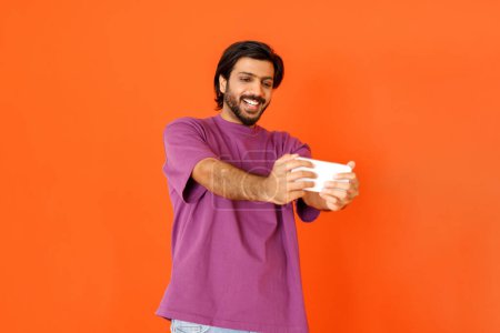 Photo for Happy cheerful smiling handsome young eastern guy in purple t-shirt taking selfie on smartphone, have online meeting video call with friends, orange studio background, copy space - Royalty Free Image