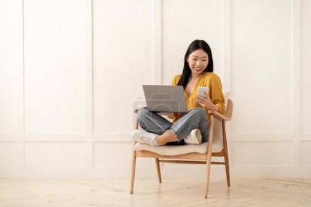 Photo for Gadget addiction. Positive cheerful pretty young asian woman freelancer working from home, female sitting in armchair, using laptop and checking messages on smartphone, copy space - Royalty Free Image