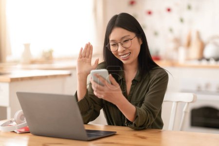 Téléchargez les photos : Joyeux jeune jolie femme asiatique portant des lunettes assis au bureau de la cuisine devant un ordinateur portable, regardant l'écran du smartphone et gestualisant, avoir un appel vidéo avec un partenaire d'affaires, copier l'espace - en image libre de droit