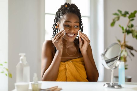 Gentle touch of freshness. Beautiful black lady cleansing skin with cotton pads, looking at mirror and smiling, woman enjoying beauty skincare routine at home, copy space