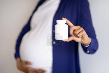 Foto de Millennial africana americana embarazada mujer con gran vientre muestra frasco de vitaminas píldoras sobre fondo de pared gris. Expectativa del niño, atención médica en el hogar, tratamiento y recomendación, publicidad y oferta - Imagen libre de derechos