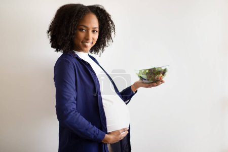 Foto de Encantada señora embarazada afroamericana milenaria con una gran ensalada de guarnición en el fondo de la pared gris. Alimentación saludable, alimentación nutricional, dieta de recomendación, embarazo infantil, atención médica en el hogar - Imagen libre de derechos