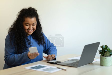 Photo for Positive cheerful curly chubby young woman in casual outfit entrepreneur working from home, sitting at desk, using laptop, calculator and smartphone, checking newest business mobile app, copy space - Royalty Free Image