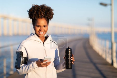 Photo for Black Sporty Female With Fitness Bottle And Smartphone Relaxing After Outdoor Training, Young African American Athlete Woman Resting After Morning Jogging, Standing On Pier And Smiling At Camera - Royalty Free Image