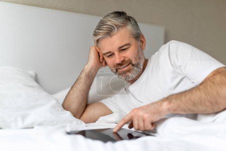 Foto de Relajado alegre guapo de pelo gris hombre de mediana edad con camiseta blanca acostado en la cama en casa, utilizando moderno pc almohadilla digital, tocando la pantalla en blanco de la mesa, desplazándose por Internet, espacio de copia - Imagen libre de derechos