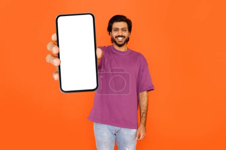 Foto de Alegre guapo barbudo joven indio mostrando enorme teléfono inteligente con pantalla blanca en blanco y sonriendo, maqueta para la oferta en línea o anuncio de aplicación móvil, aislado en fondo de estudio naranja, espacio de copia - Imagen libre de derechos