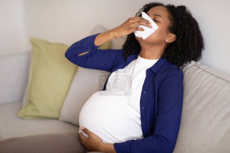 Foto de Infeliz milenaria afroamericana embarazada con gran vientre sufre de enfermedad y problemas de salud, se sopla la nariz en una servilleta. esperando un bebé maternidad y presión, gripe y resfriados - Imagen libre de derechos