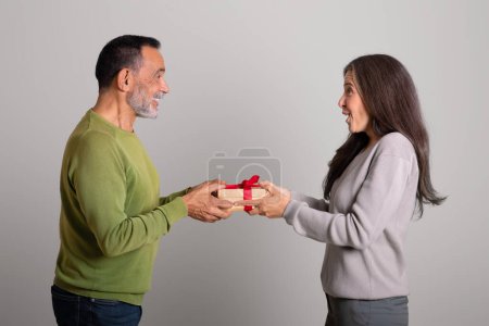 Photo for Cheerful caucasian elderly man gives gift box, congratulations to lady on gray studio background. Birthday greeting, anniversary surprise, relationship and love, celebrating holiday together - Royalty Free Image