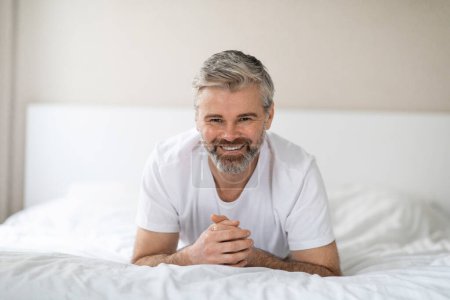 Téléchargez les photos : Portrait d'un heureux homme d'âge moyen aux cheveux gris et perlé portant un pyjama couché sur le lit dans une chambre blanche confortable à la maison, souriant à la caméra, profitant d'une bonne matinée après un bon sommeil - en image libre de droit