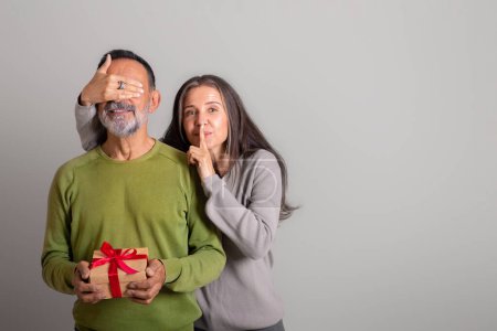Photo for Glad caucasian elderly lady closes eyes to husband, make shh sign, gives gift, congratulations on holiday on gray studio background. Birthday greeting, anniversary, surprise, relationship and love - Royalty Free Image