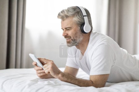 Téléchargez les photos : Relaxé heureux bel homme d'âge moyen portant un pyjama couché sur le lit à la maison, en utilisant un smartphone et un casque sans fil flambant neuf, avoir un chat vidéo, jeux en ligne, espace de copie, vue latérale - en image libre de droit