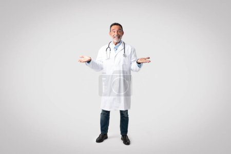 Photo for Glad european senior doctor in white coat with stethoscope spreads arms to sides, holds empty space on gray background. Choice, professional advice ,medical care, exam, ad and offer - Royalty Free Image