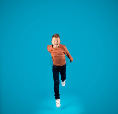 Téléchargez les photos : Preteen Boy sautant dans les airs et regardant la caméra, en mouvement prise de vue de gai caucasien entre mâle enfant duper et s'amuser sur fond bleu dans Studio, pleine longueur, Copier l'espace - en image libre de droit