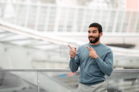 Photo for Check This Out. Cheerful Middle Eastern Man Pointing Fingers Aside At Copy Space Advertising Great Travel Offer Posing At Modern Airport. Look There, Cheap Tickets Advertisement - Royalty Free Image