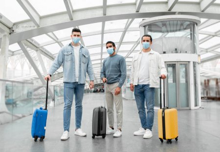 Foto de Tres viajeros hombres que usan máscaras faciales para la protección contra el virus de pie con maletas de viaje en la moderna terminal de salida del aeropuerto en interiores. Concepto de Transporte Seguro. Disparo de longitud completa - Imagen libre de derechos