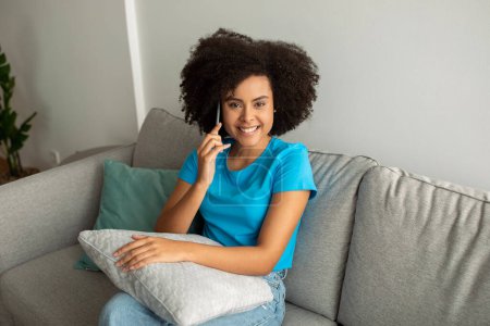 Photo for Good news, gossip. Glad pretty young black curly woman calling by smartphone, talking to friend on couch in living room interior. Spare time, communication at work at home - Royalty Free Image