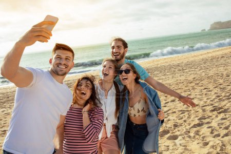 Foto de Felices amigos milenarios europeos y árabes se divierten, tomando selfie en el teléfono inteligente, disfrutar del tiempo libre en la playa del océano. Aplicación para blog y red social, fiesta al aire libre, vacaciones y viaje - Imagen libre de derechos