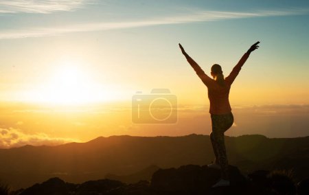 Foto de Vista posterior de la mujer turista de pie en la cima de la montaña levantando brazos mirando la hermosa puesta de sol por encima de las nubes. Silueta del Exitoso Senderista. Objetivos de viaje y concepto de logro - Imagen libre de derechos