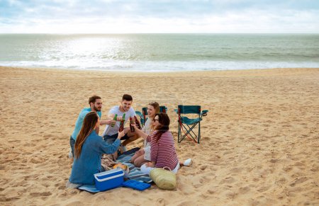Foto de Felices amigos milenarios europeos y árabes se divierten, disfrutan del tiempo libre, animan botellas, hacen picnic en la playa del océano. Vacaciones juntos, fiesta al aire libre, vacaciones en equipo en verano - Imagen libre de derechos