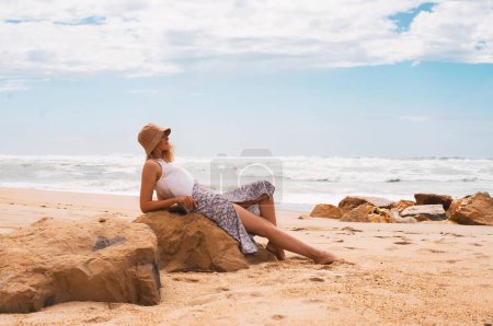Téléchargez les photos : Voyage de vacances. Femme relaxant et bronzant sur la plage près de l'océan, regardant l'eau et les vagues assis sur la pierre profitant d'une journée ensoleillée au bord de la mer en plein air. Voyageuse se reposant sur la côte de la mer - en image libre de droit