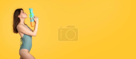 Foto de Joven mujer europea fresca en ropa de baño sosteniendo pistola de agua, divirtiéndose en el fondo del estudio amarillo, panorama con espacio para copiar, vista lateral. Actividad acuática durante las vacaciones de verano - Imagen libre de derechos