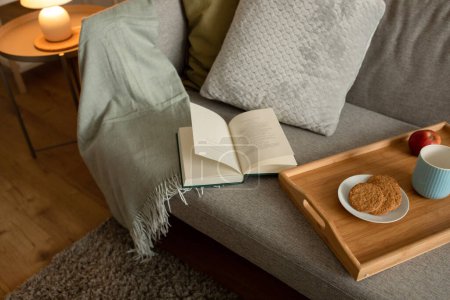 Photo for Book, tray with food and coffee cup on sofa in living room interior at evening, top view. Hobbies, literature in free time, cozy, comfort at home, study, education and fairytale, nobody - Royalty Free Image