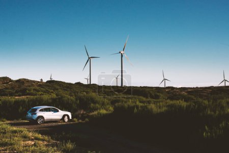 Photo for Electric Car Parked In Field Near High Wind Turbines Generating Green Electiricity Outside. Modern Windmills Farm Background With New Automobile. Renewable Energy Sources Concept - Royalty Free Image