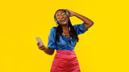 Foto de Aplicación de música. Mujer afroamericana con estilo con teléfono escuchando canciones en línea presionando auriculares para oír, disfrutando y relajándose con los ojos cerrados sobre fondo amarillo del estudio. Panorama - Imagen libre de derechos