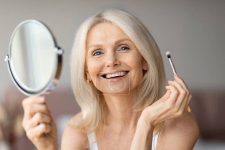 Téléchargez les photos : Heureuse dame européenne âgée utilisant brosse et fards à paupières tenant miroir pour le maquillage nu, souriant à la caméra. Cosmétiques anti-âge, soins de beauté, procédures de routine, publicité et offre - en image libre de droit