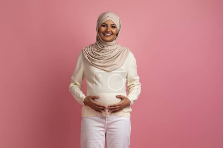 Foto de Hermosa mujer musulmana embarazada abrazando tiernamente el vientre mientras posaba contra el fondo del estudio rosa, feliz madre árabe embarazada acariciando el golpe del bebé y sonriendo en la cámara, espacio de copia - Imagen libre de derechos
