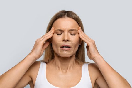 Blonde middle aged woman with closed eyes rubbing temples on grey studio background, experiencing strong headache, suffering from migraine, closeup photo, copy space