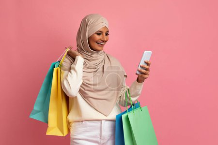 Téléchargez les photos : Concept d'achat en ligne. Femme musulmane souriante utilisant un téléphone intelligent et tenant des sacs Shopper, Happy Islamic Female In Hijab bénéficiant d'une application mobile pour les achats sur Internet, posant sur fond rose - en image libre de droit