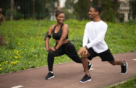 Téléchargez les photos : Un jeune couple afro-américain positif en tenue de sport faisant de l'exercice de jambes, s'étirant et se réchauffant dans le parc. Entraînement ensemble en ville, mode de vie actif et perte de poids, soins du corps en plein air - en image libre de droit
