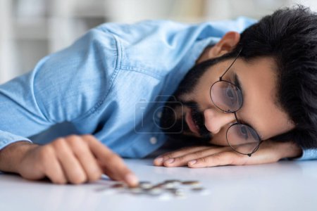 Photo for Financial Problems. Depressed Indian Man Counting Coins Lying On Table, Upset Young Eastern Male Suffering Money Crisis, Sad Guy Having Troubles With Budget Planning, Closeup Shot, Selective Focus - Royalty Free Image
