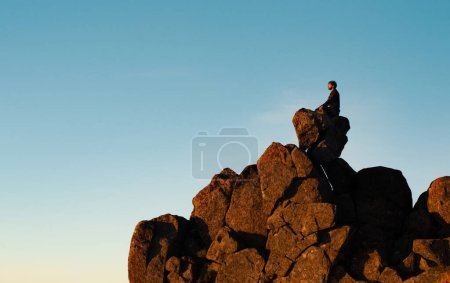 Foto de Vista lateral del hombre pacífico sentado en la cima del acantilado de roca meditando y relajándose con los ojos cerrados afuera. Tipo viajero posando en posición de loto haciendo yoga al aire libre en las montañas. Mindfulness - Imagen libre de derechos