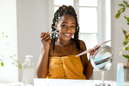 Photo for Happy black woman applying makeup at home, beautiful lady sitting in front of mirror, holding eyeshadow palette and brush, putting cosmetics on her face, copy space. Home beauty studio concept - Royalty Free Image