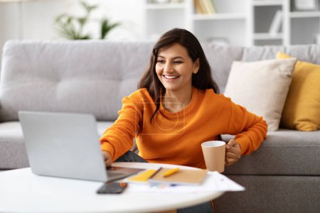 Photo for Happy cheerful beautiful millennial eastern lady working from home, sitting on floor next to couch, using modern pc laptop, drinking coffee, copy space. Remote job, freelance concept - Royalty Free Image
