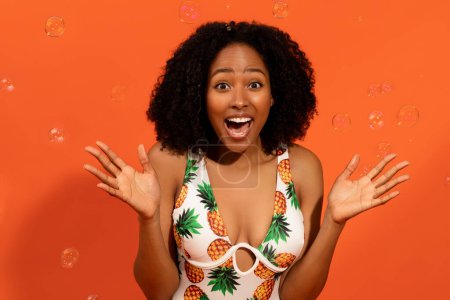 Photo for Emotional happy attractive millennial african american woman wearing fashionable summer swimsuit posing among water bubbles on orange background, gesturing and exclaiming, enjoying vacation, closeup - Royalty Free Image