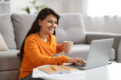 Happy pretty young long-haired indian woman in casual outfit freelancer sitting on floor by couch, typing on pc laptop keyboard and smiling, drinking coffee, working from home, copy space magic mug #656528402