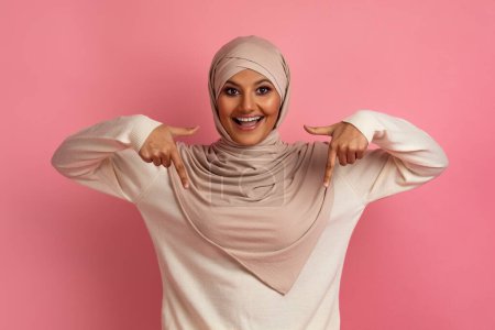 Photo for Look There. Excited muslim woman in hijab pointing down with two fingers, smiling islamic woman in headscarf demonstrating copy space for your design or advertisement, standing over pink background - Royalty Free Image
