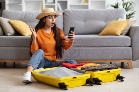 Photo for Shocked pretty young eastern woman wearing comfy casual outfit and summer hat traveller sitting on floor, packing suitcase, looking at phone screen and gesturing, late for flight, home interior - Royalty Free Image