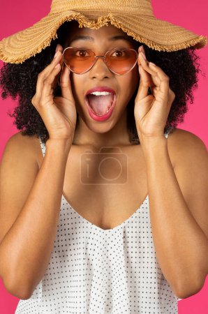 Foto de Alegre señora rizada afroamericana milenaria sorprendida con boca abierta, sombrero y gafas de sol aisladas sobre fondo rosa, estudio, vertical. Diversión de verano, relax y vacaciones - Imagen libre de derechos