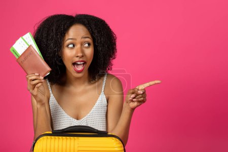 Photo for Glad surprised millennial black curly lady with passport, tickets and suitcase pointing finger at empty space isolated on pink background, studio. Summer vacation and travel, ad and offer - Royalty Free Image