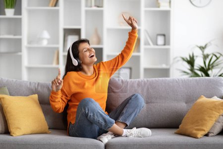 Joyful beautiful millennial eastern lady wearing casual outfit sitting on couch at home, listening to music with closed eyes, dancing and singing, using wireless headphones, copy space