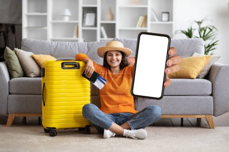 Photo for Cheery happy young eastern woman wearing casual comfy outfit and straw hat sitting on floor by suitcase at home, tourist showing phone with blank screen, recommending nice mobile app, mockup - Royalty Free Image