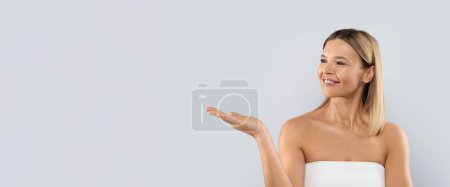 Téléchargez les photos : Jolie jolie blonde d'âge moyen enveloppée dans une serviette montrant quelque chose d'invisible sur sa paume et souriant, isolée sur fond de studio, panorama. Soins du visage cosmétique concept - en image libre de droit