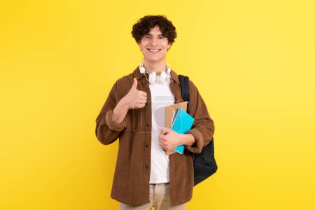 Photo for Great Course. Happy Young Man Gesturing Thumbs Up Expressing Approval And Optimism, Recommending His University Study Program, Posing With Backpack And Textbooks On Yellow Studio Background - Royalty Free Image